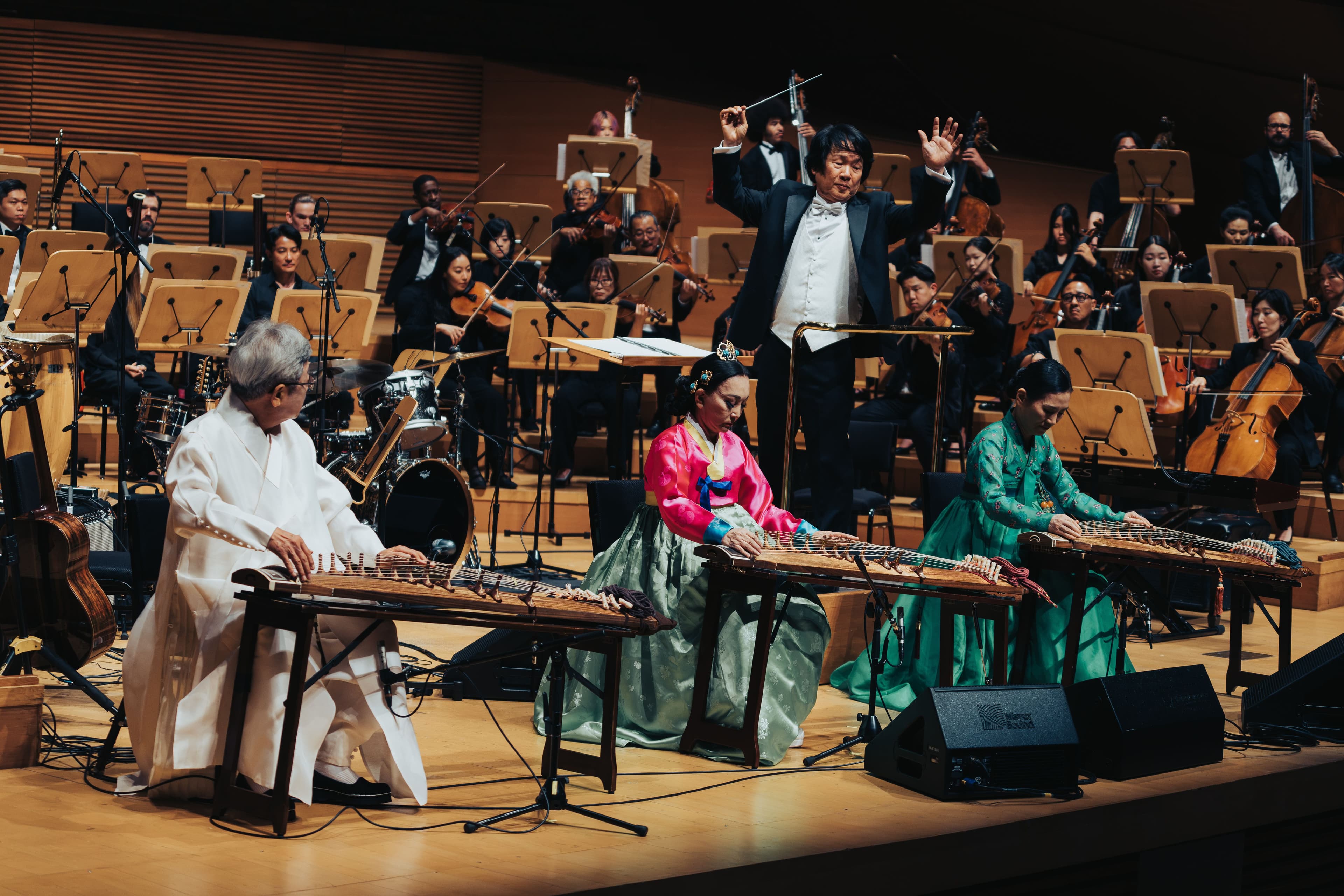 Three musicians in traditional Korean clothing play gayageum at the front of the stage while a conductor leads the orchestra behind them.