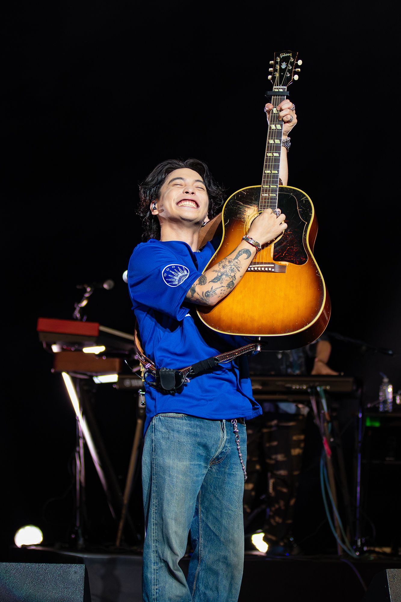 Yuuri smiles with his eyes closed while lifting an acoustic guitar overhead in a bright blue Wiltern jersey.