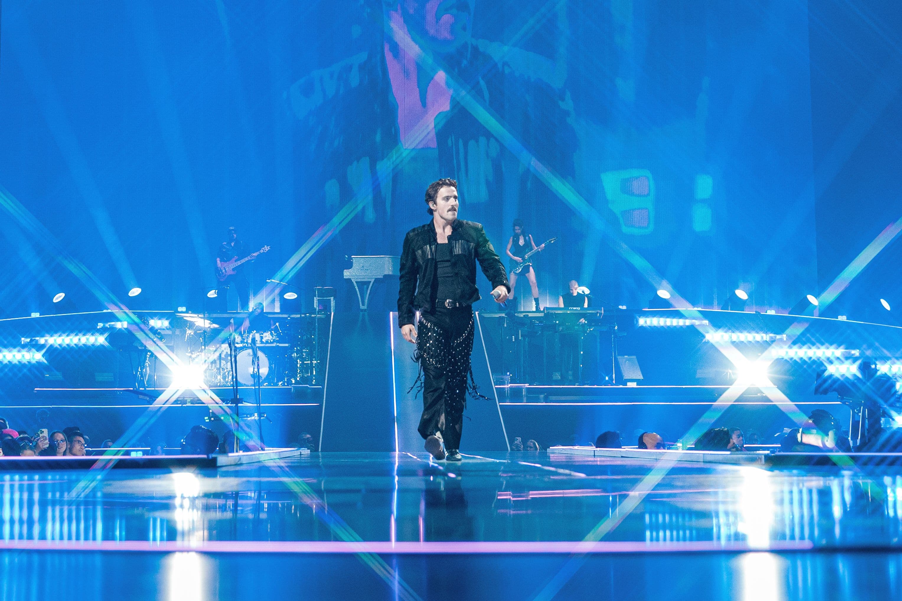 Benson Boone walks down the illuminated center runway at Crypto.com Arena as blue stage lights and lasers fill the arena behind him.