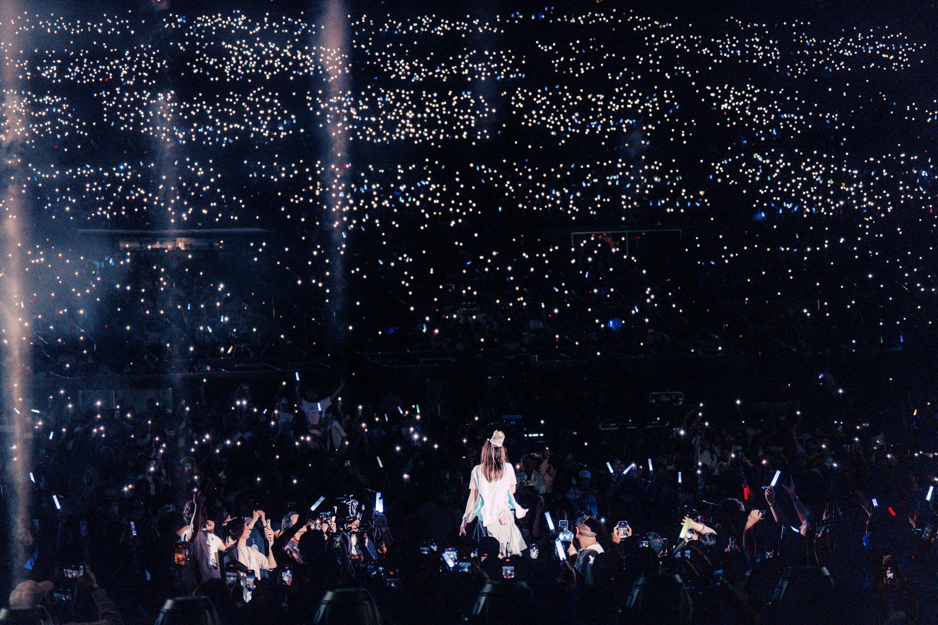 YOASOBI vocalist ikura stands among the crowd at the Hollywood Bowl as thousands of fans illuminate the amphitheater with phone lights and light sticks.