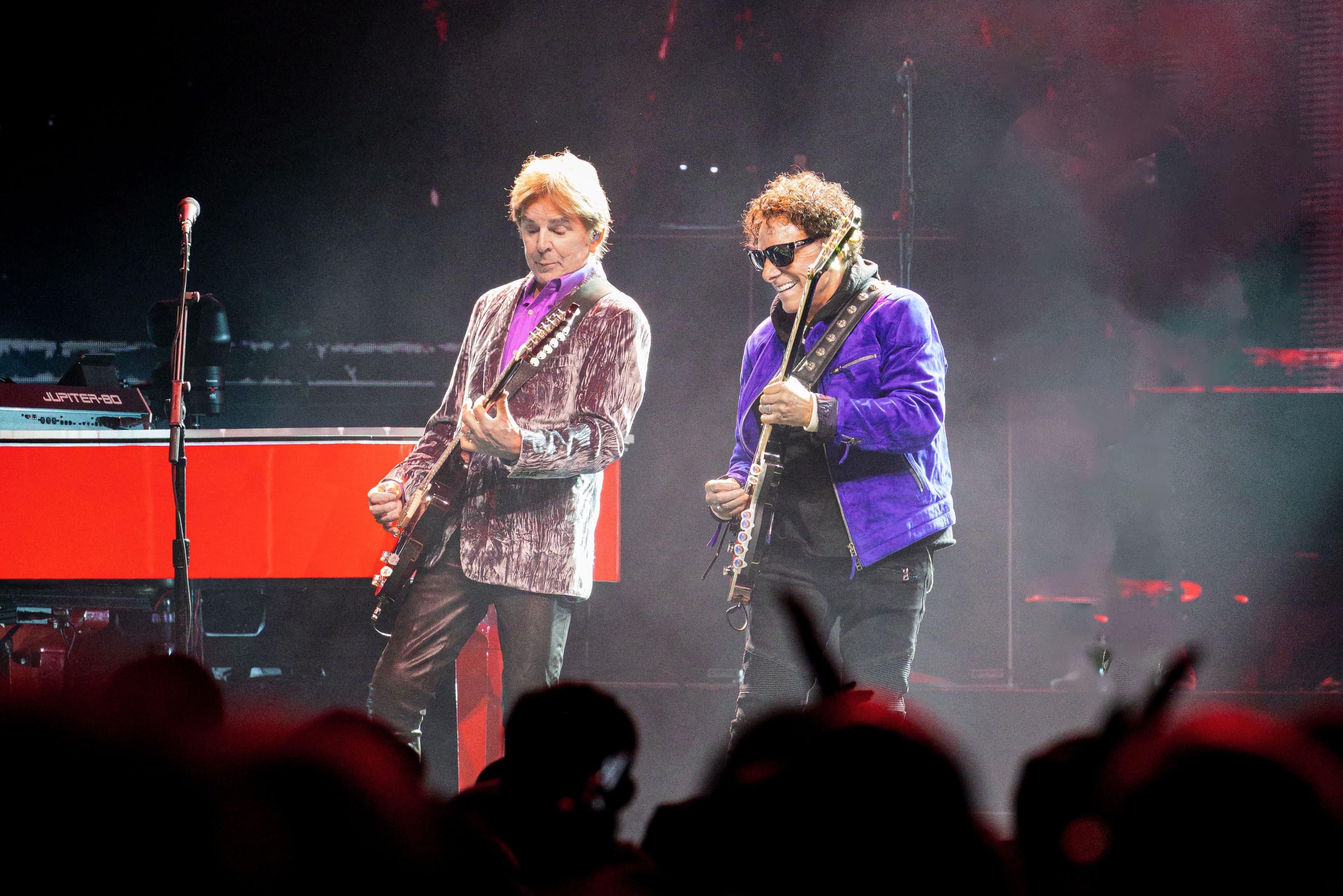 Journey performs on a large red-lit arena stage surrounded by video screens as the audience watches from the foreground.