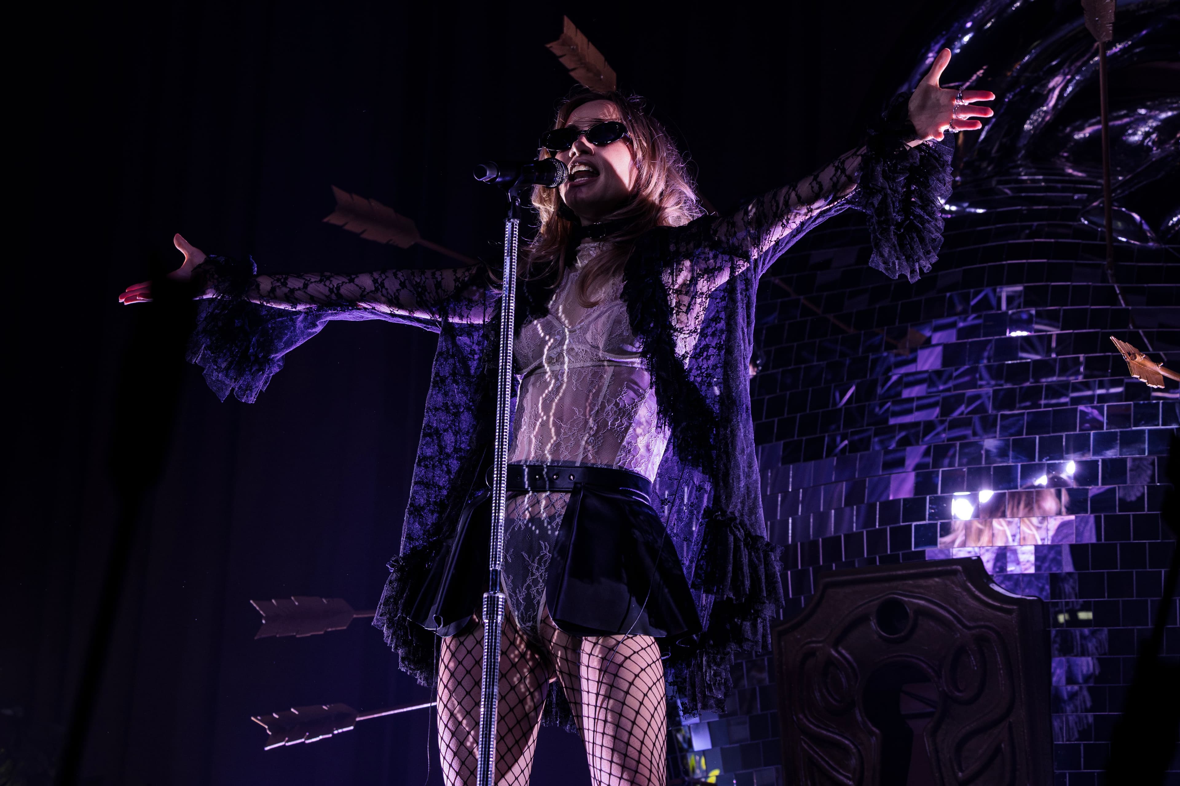 Suki Waterhouse performs with both arms stretched outward in front of a mirrored heart pierced with arrows.