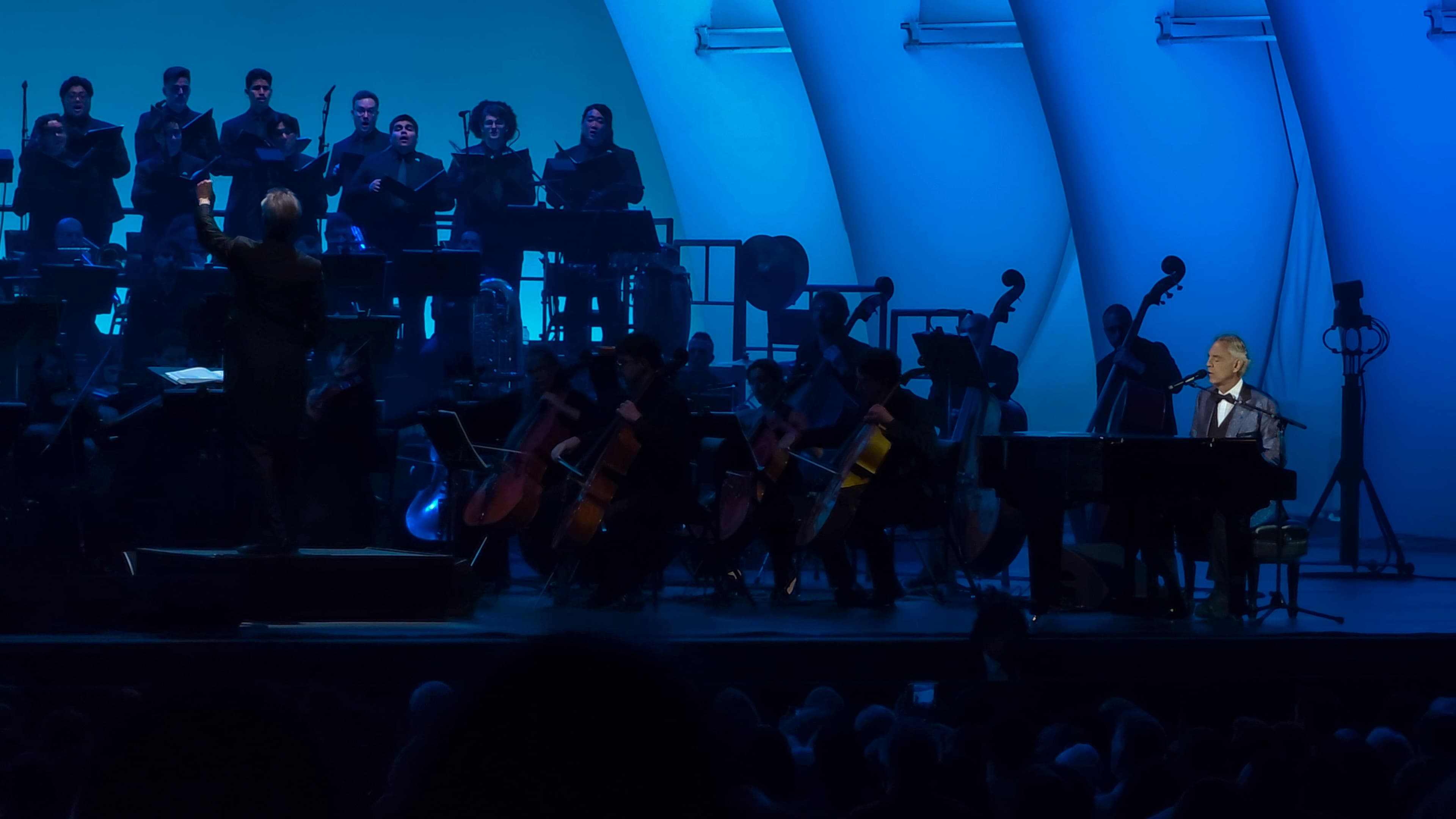 Andrea Bocelli sings from a grand piano at the Hollywood Bowl as conductor Steven Mercurio leads a full orchestra and choir under blue stage lighting.