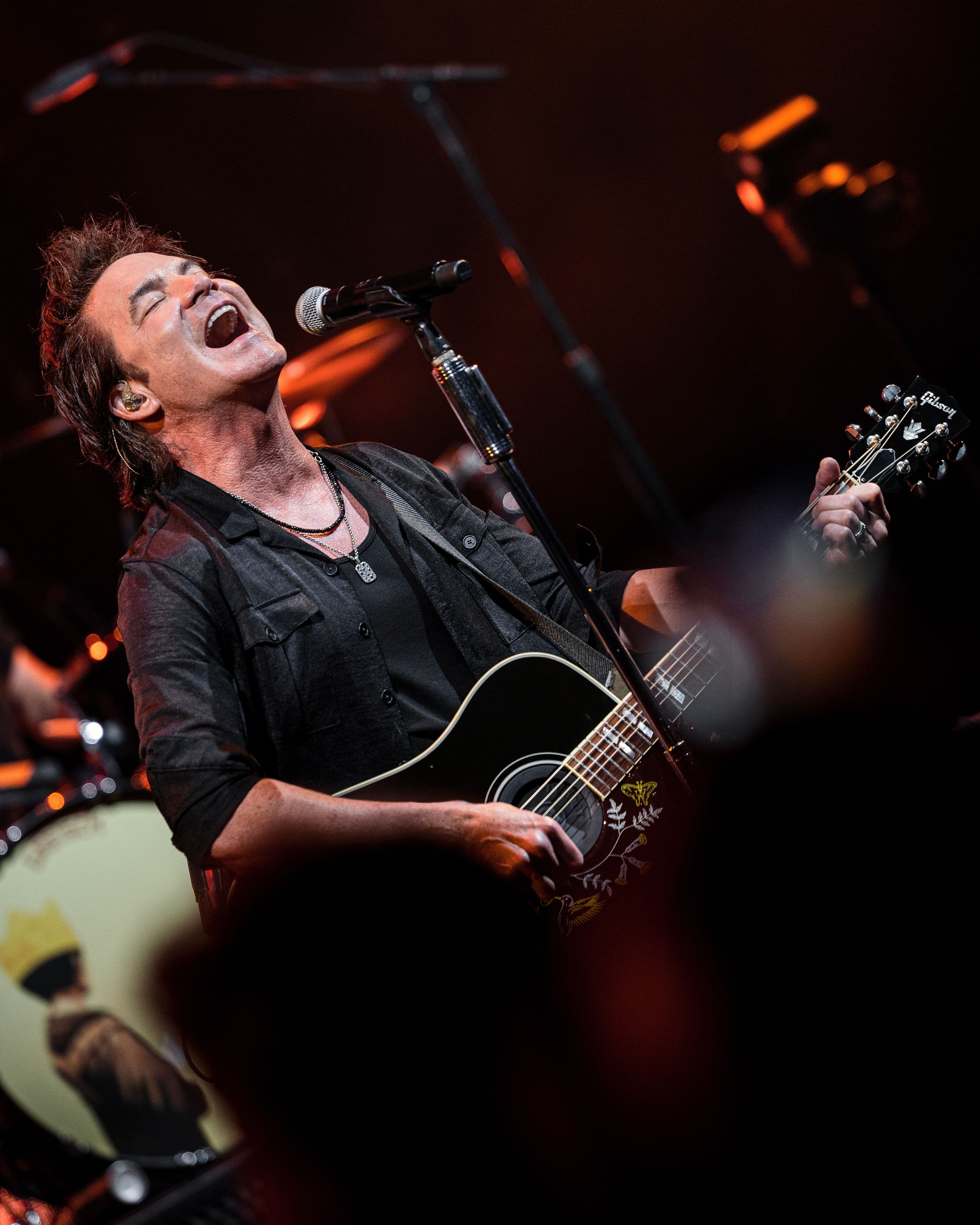 Train’s vocalist sings into a microphone while playing an acoustic guitar under warm stage lighting at the Greek Theatre.