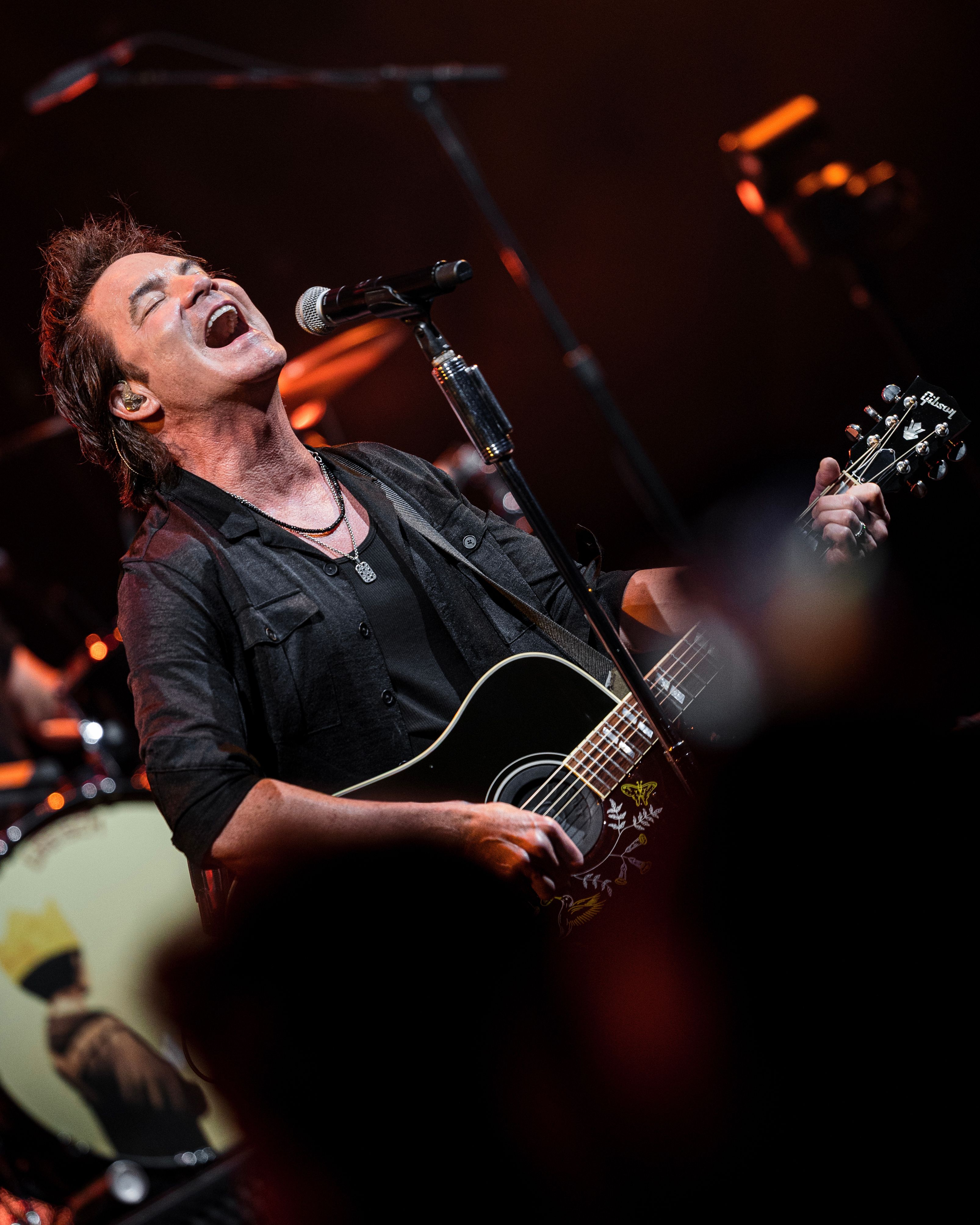 Train’s vocalist sings into a microphone while playing an acoustic guitar under warm stage lighting at the Greek Theatre.