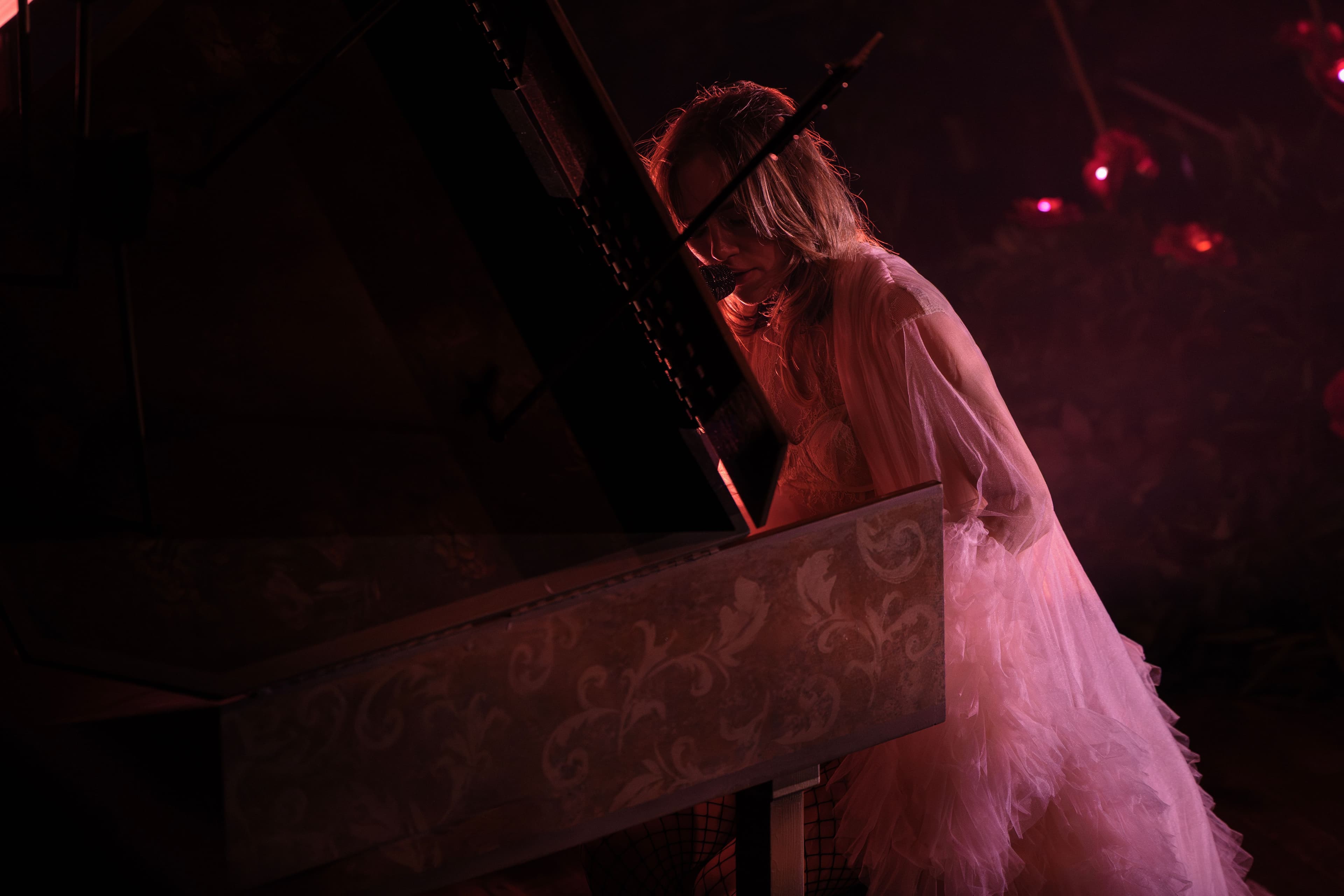 Suki Waterhouse sits at a piano under deep red lighting while wearing a flowing pale pink robe.