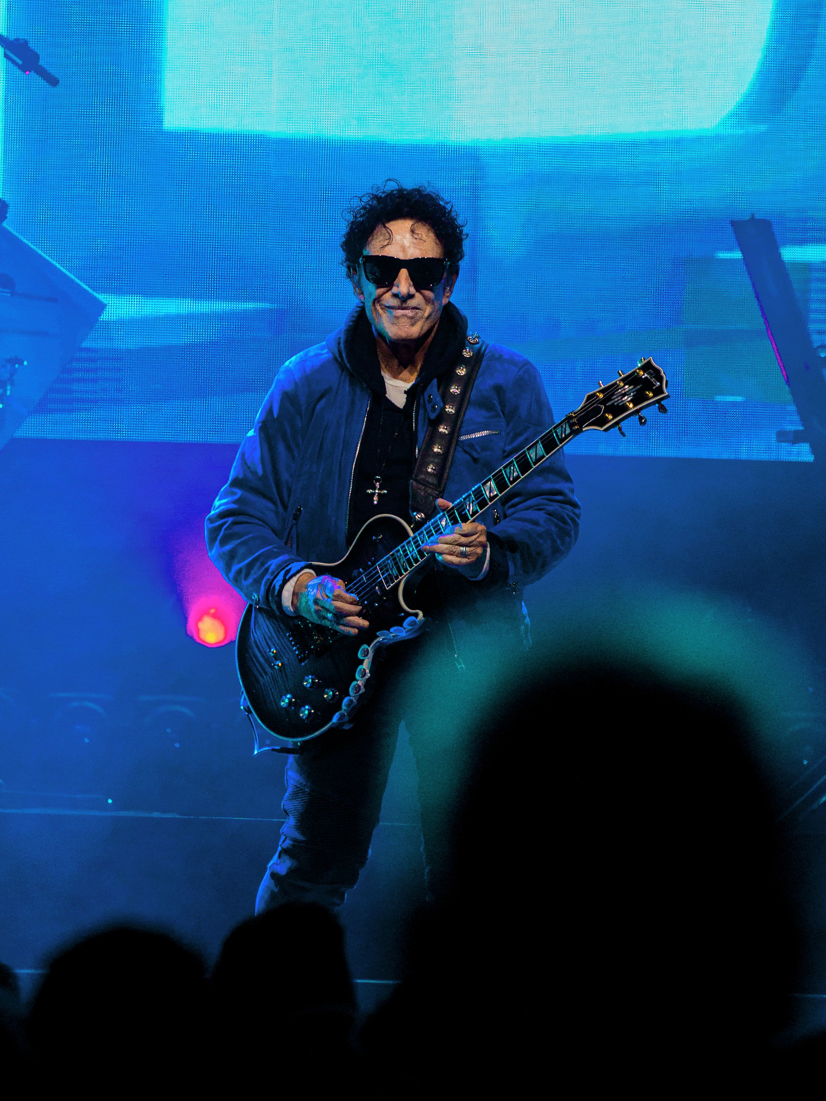 A Journey guitarist wearing sunglasses performs under saturated blue lighting with the audience silhouetted in the foreground.