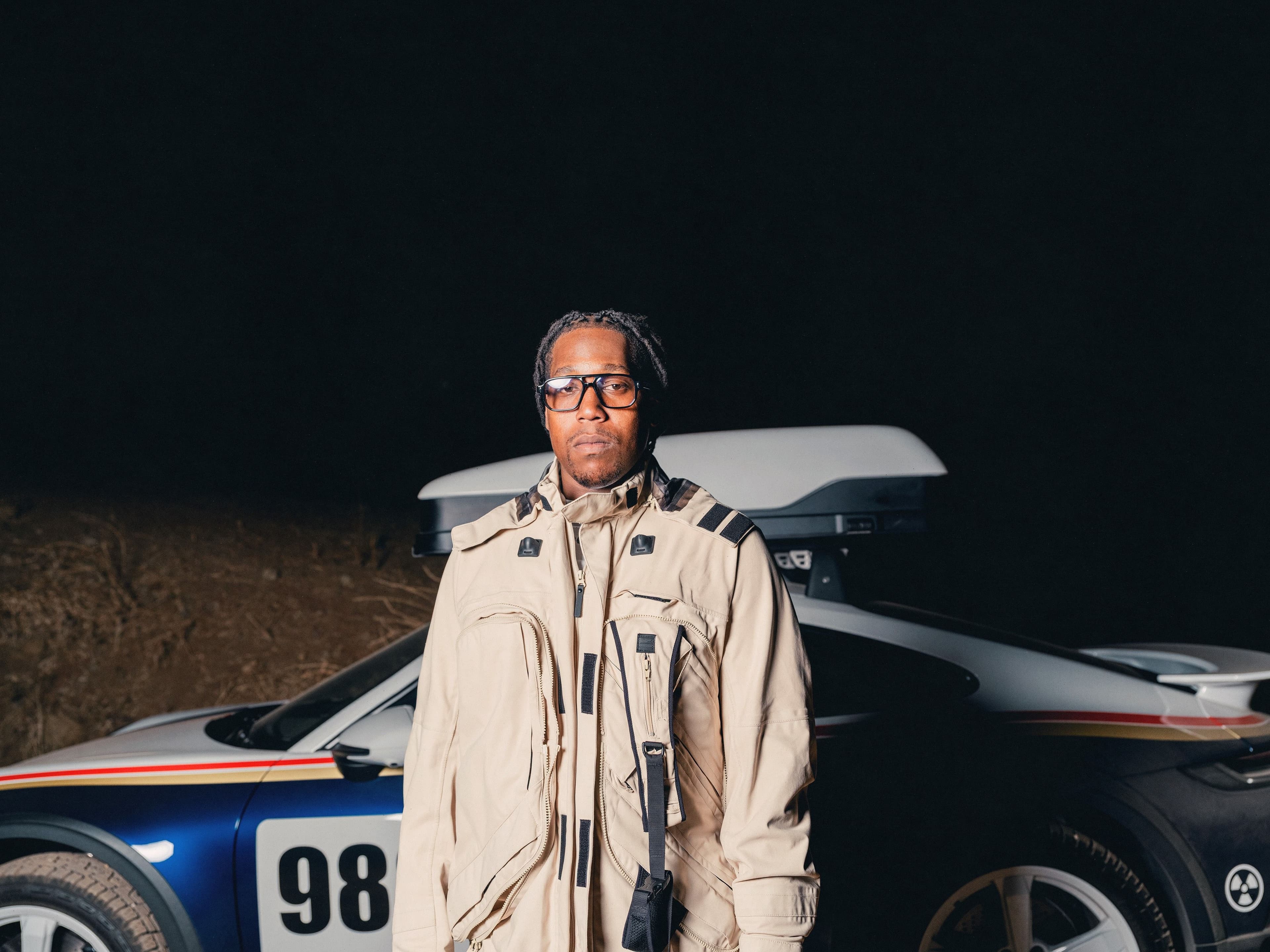 Don Toliver stands in front of a sports car at night wearing a beige technical jacket and dark glasses.
