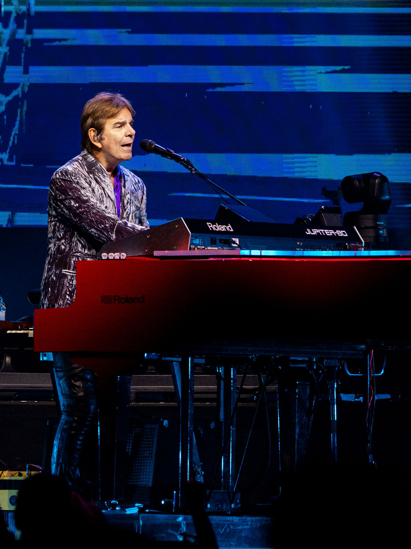 A Journey keyboardist sings while performing behind a large red keyboard under blue stage lighting.