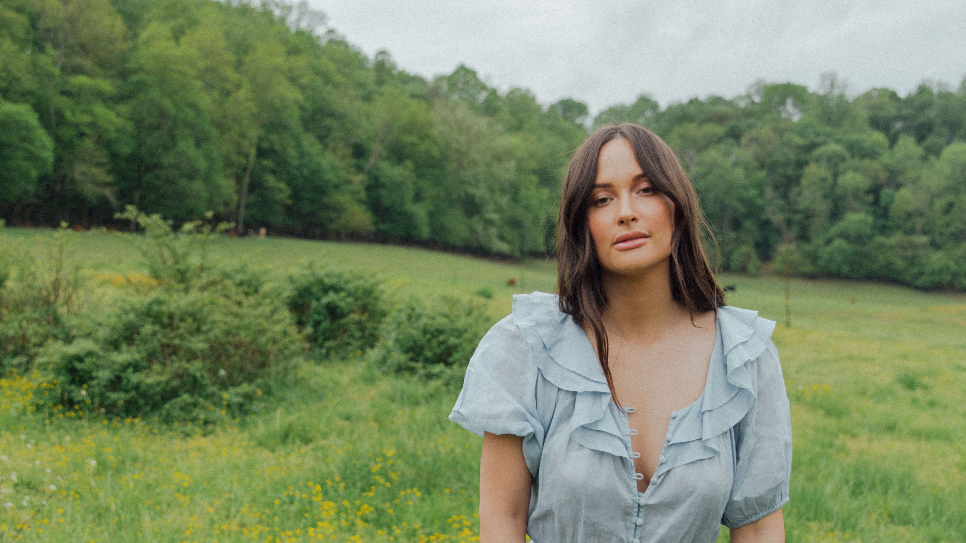 Kacey Musgraves stands in a green meadow wearing a pale blue dress.