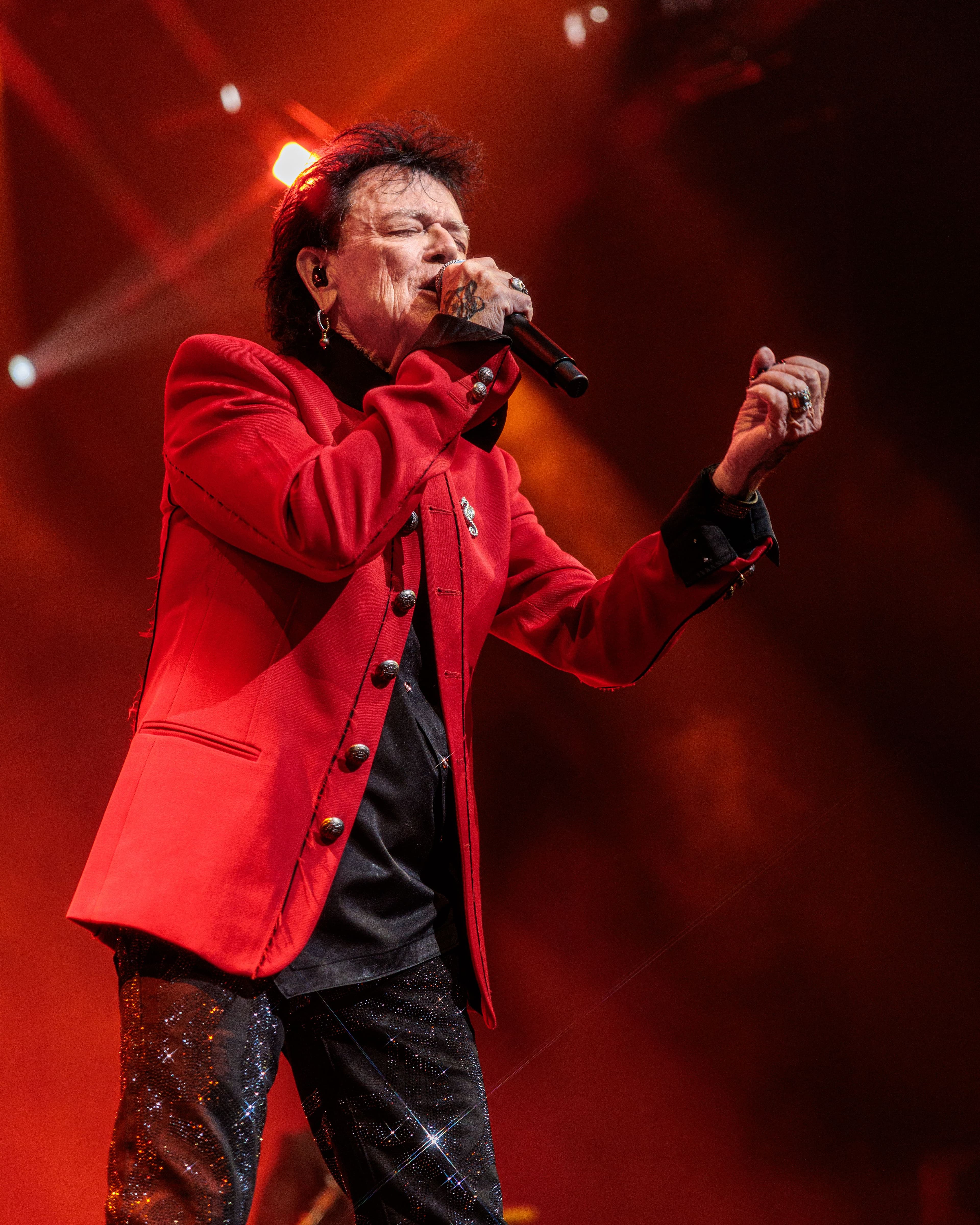 Russell Hitchcock sings into a handheld microphone wearing a bright red jacket under warm stage lighting.