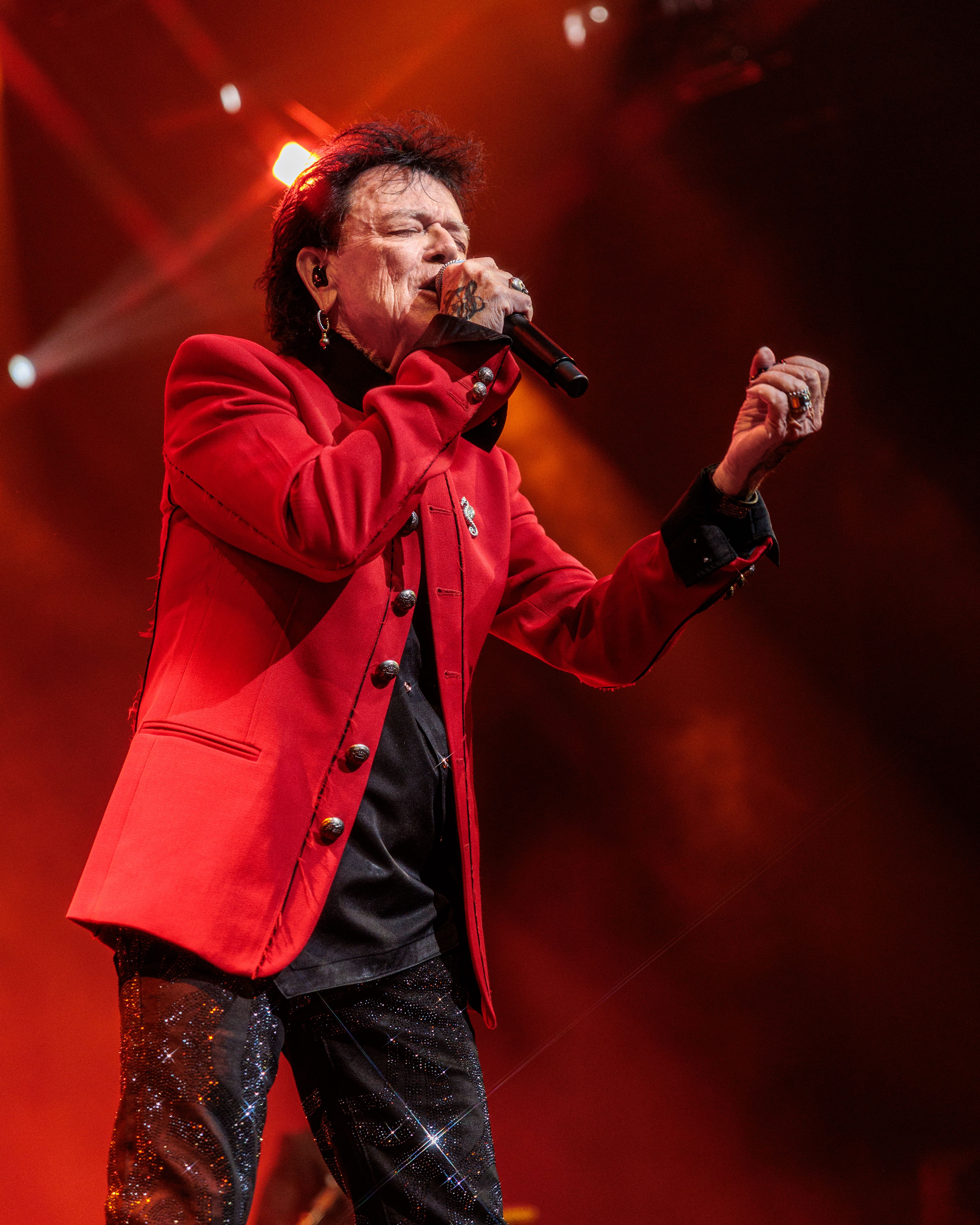 Russell Hitchcock sings into a handheld microphone wearing a bright red jacket under warm stage lighting.