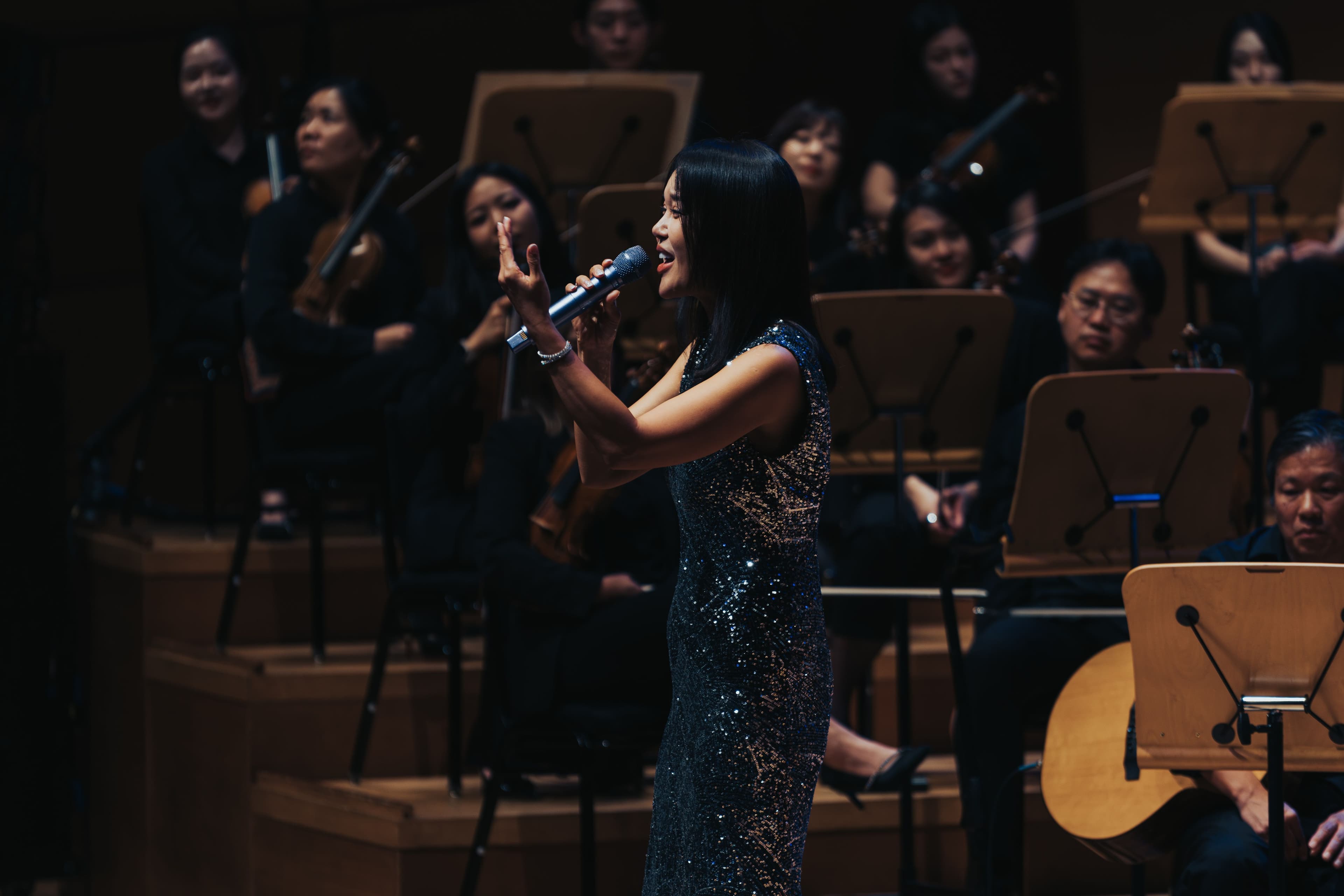 Baek Ji-young sings into a handheld microphone in a sparkling black gown with members of the orchestra seated behind her