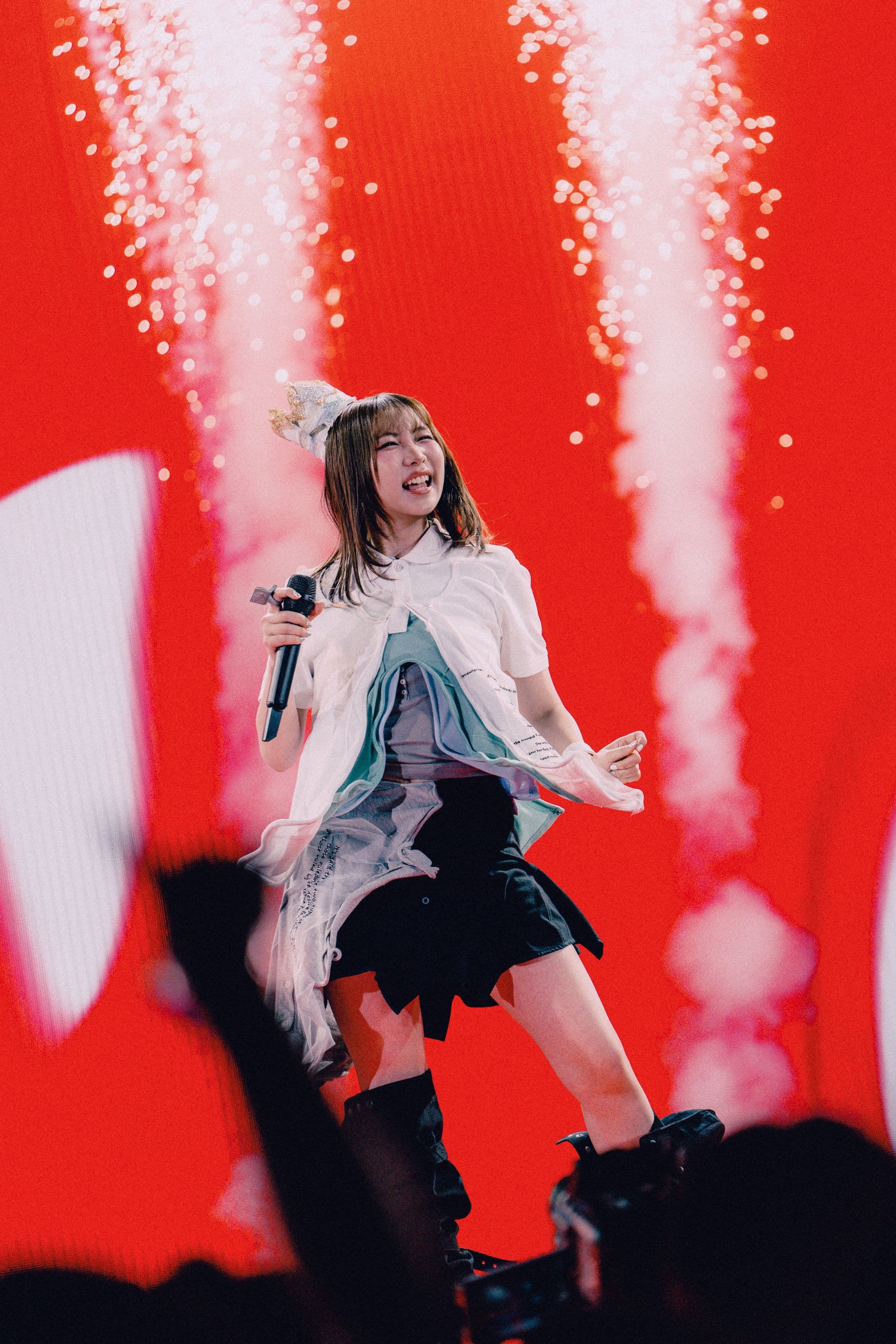 YOASOBI vocalist ikura smiles onstage at the Hollywood Bowl as bright red visuals and bursts of stage effects rise behind her.