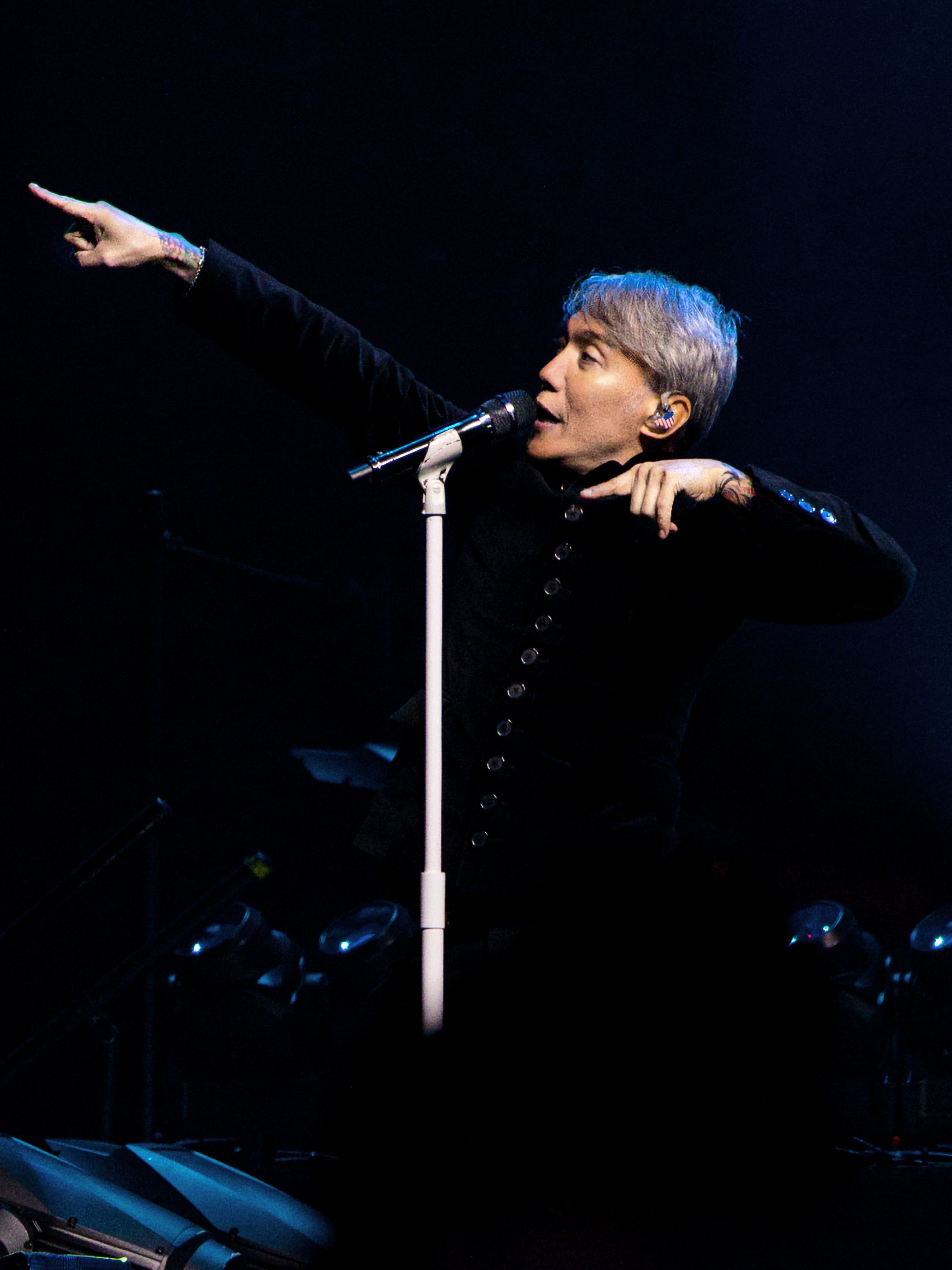 Journey’s vocalist points toward the audience while singing beside a white microphone stand under blue stage lighting.