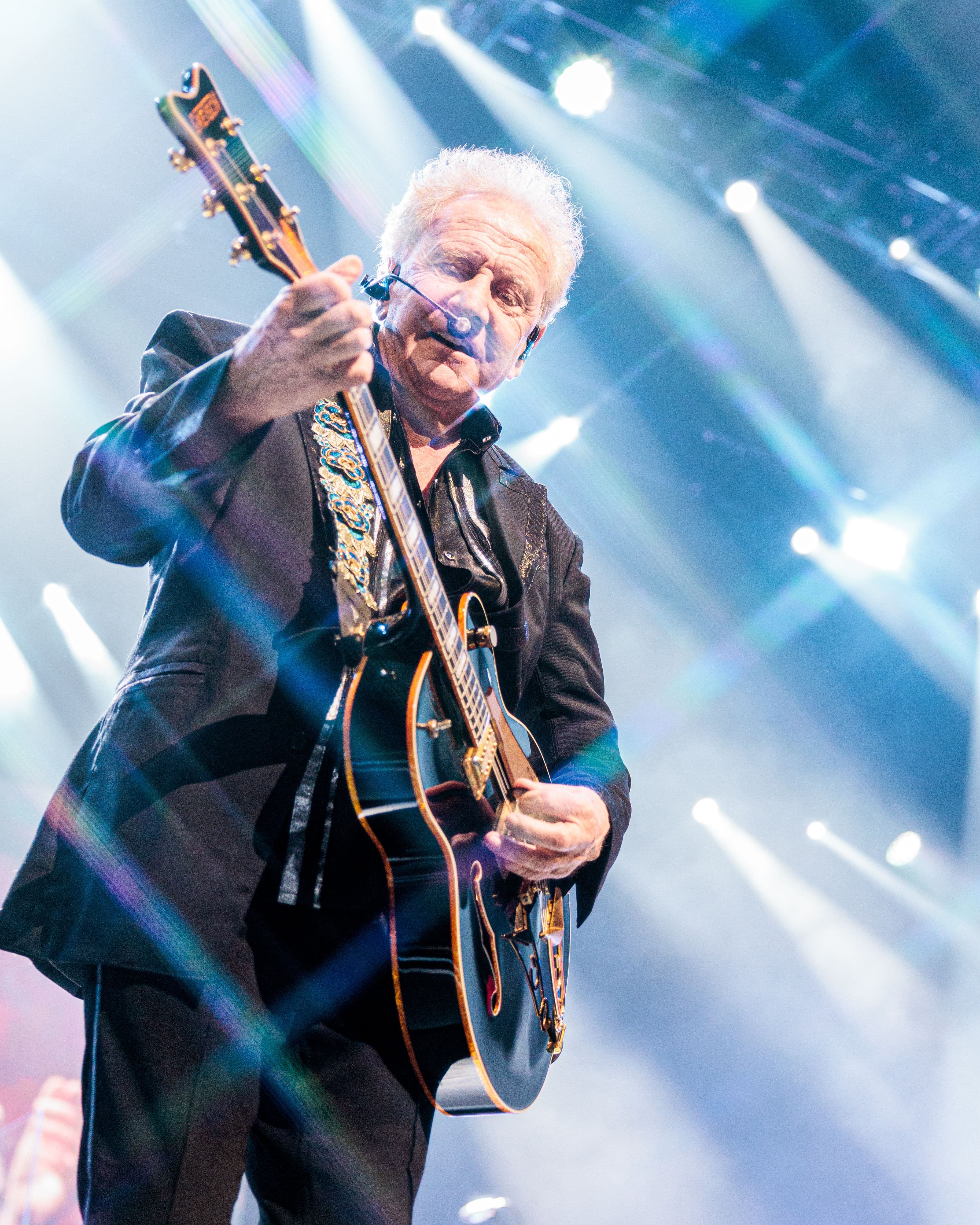 Graham Russell plays a black guitar from the front of the stage as bright white and blue lights shine behind him.