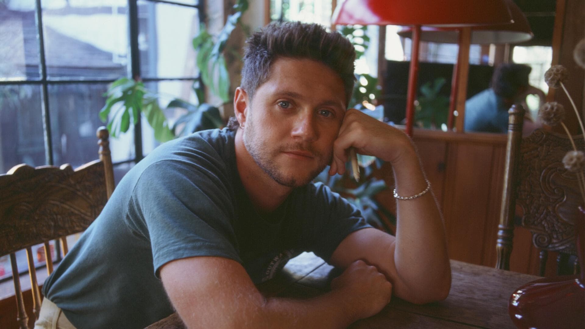 Niall Horan leans on a wooden table in a warmly lit, plant-filled room.