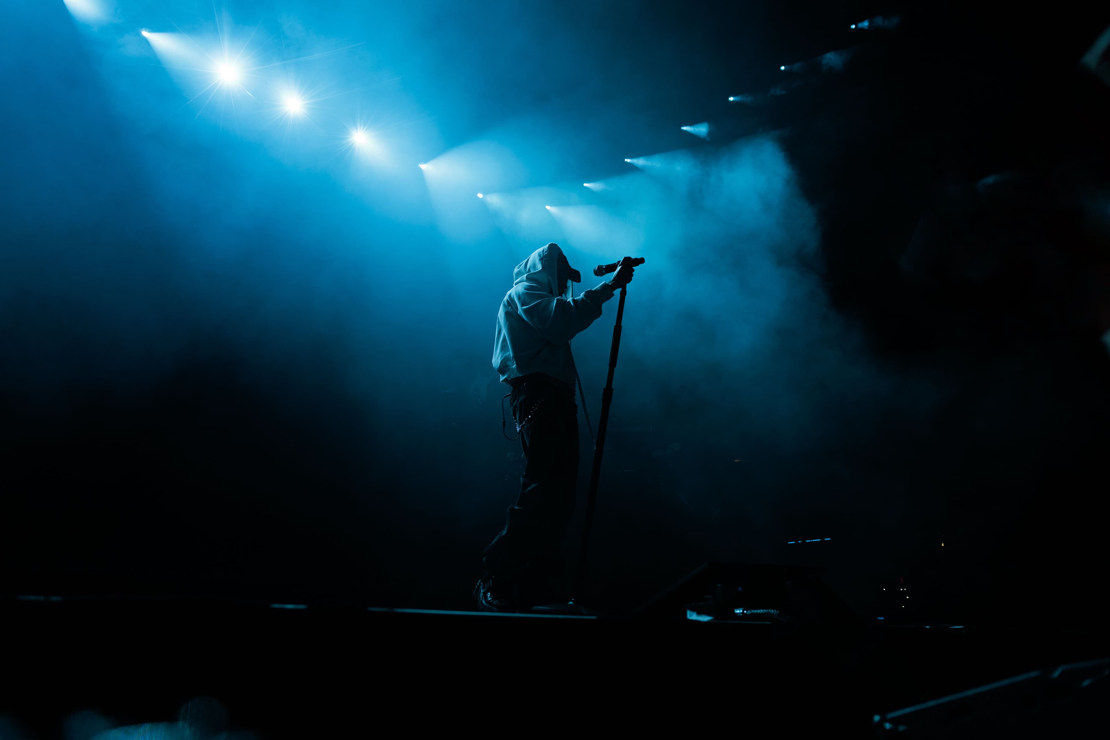 keshi stands silhouetted beneath blue spotlights and stage haze while holding a microphone stand at the Kia Forum.