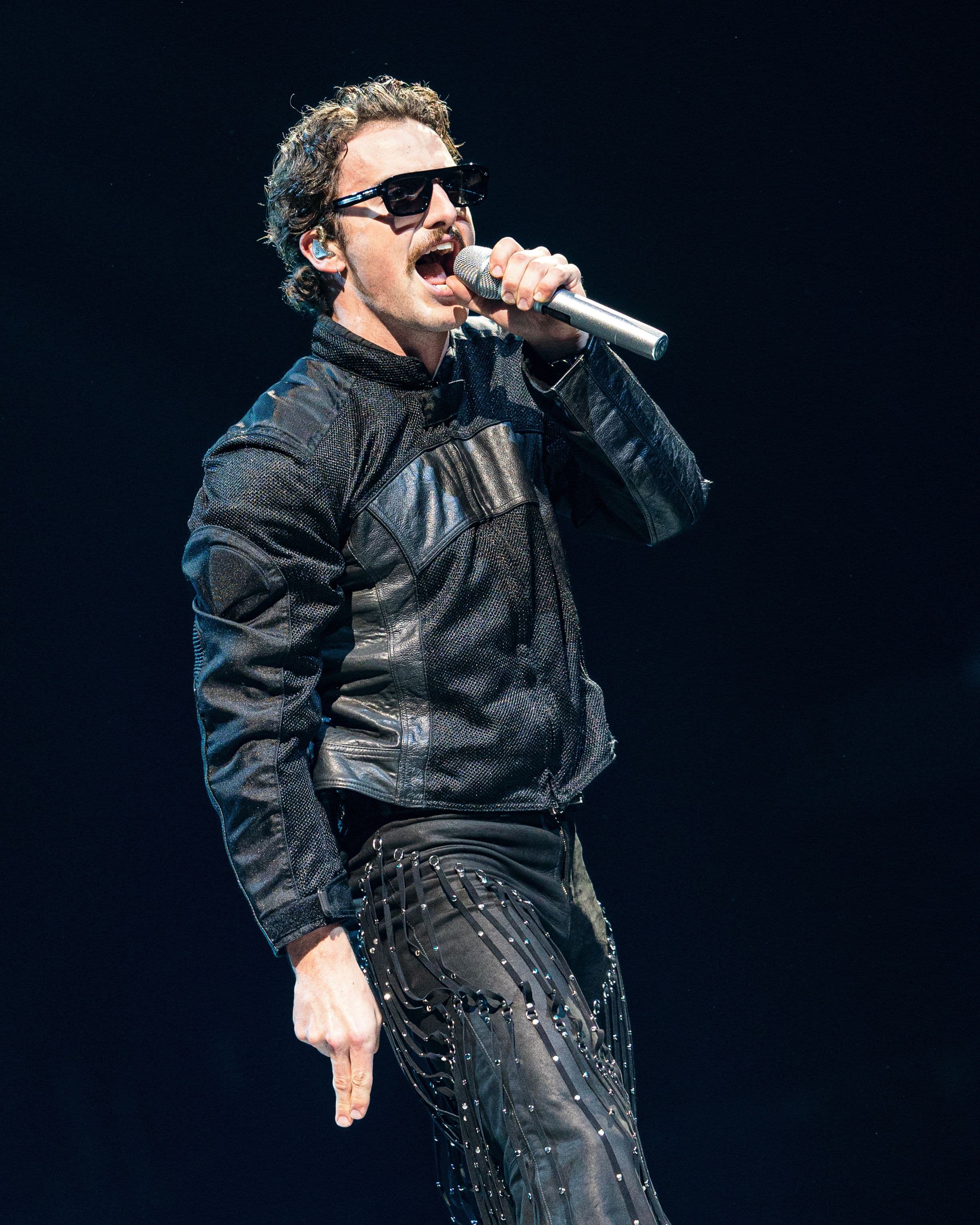 Benson Boone sings into a silver microphone wearing sunglasses, a black leather jacket and embellished black pants at Crypto.com Arena.