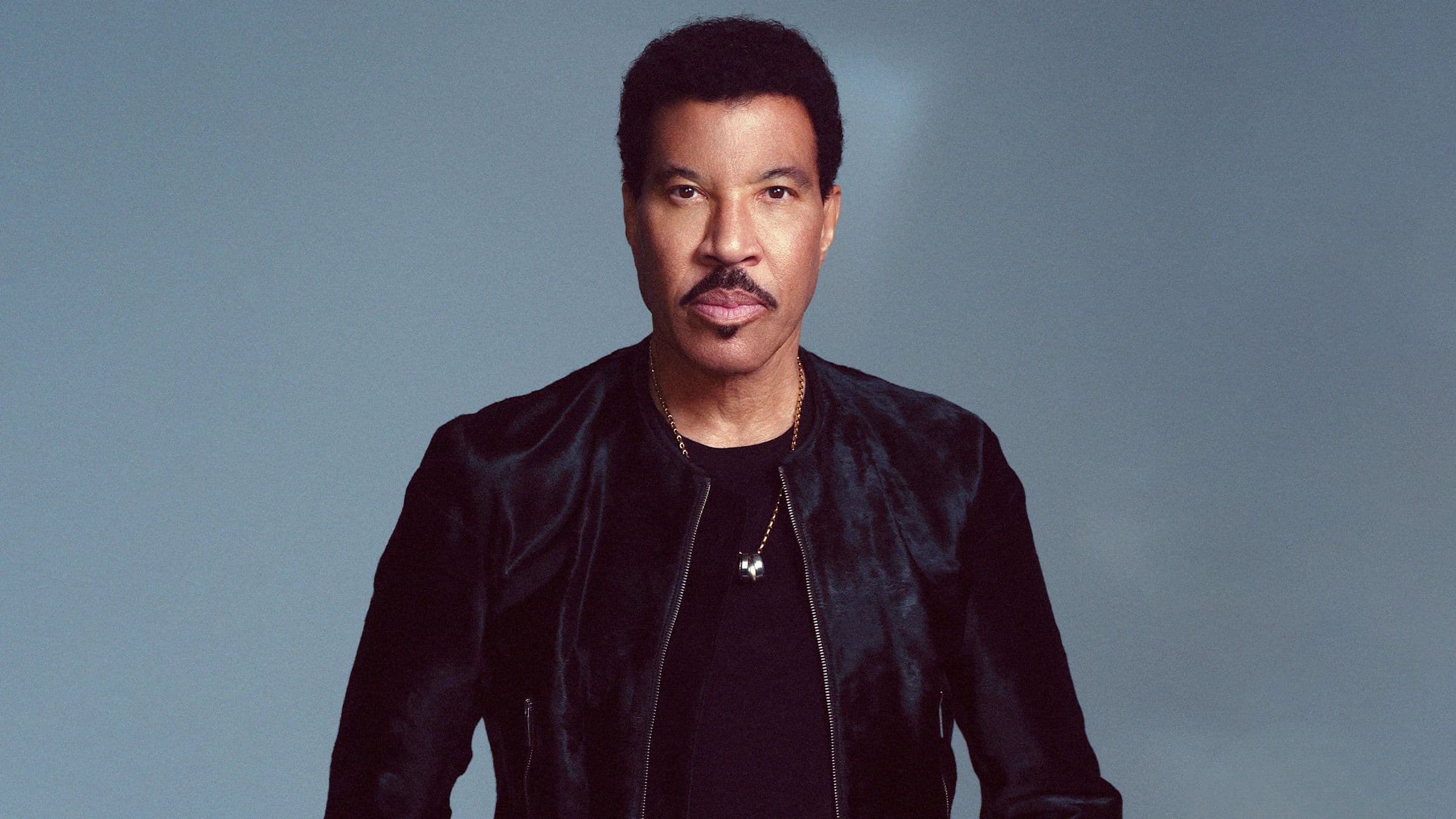 Lionel Richie poses in a black jacket against a gray studio backdrop.