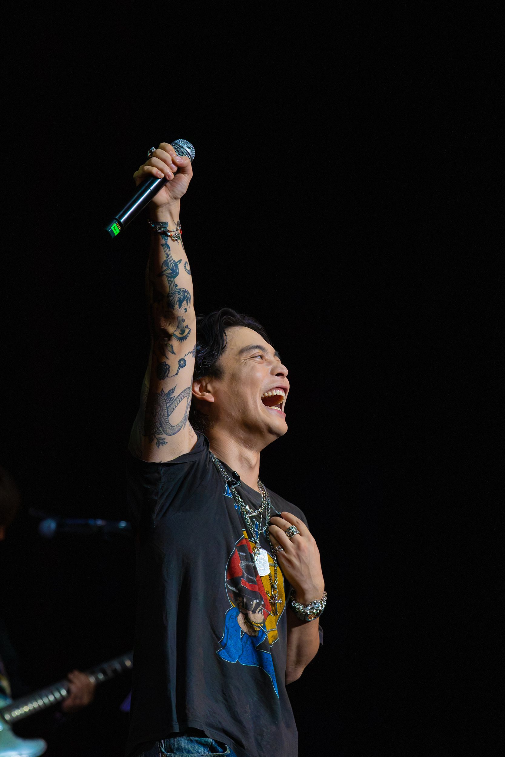 Yuuri smiles and raises a handheld microphone above his head against the dark stage at The Wiltern.